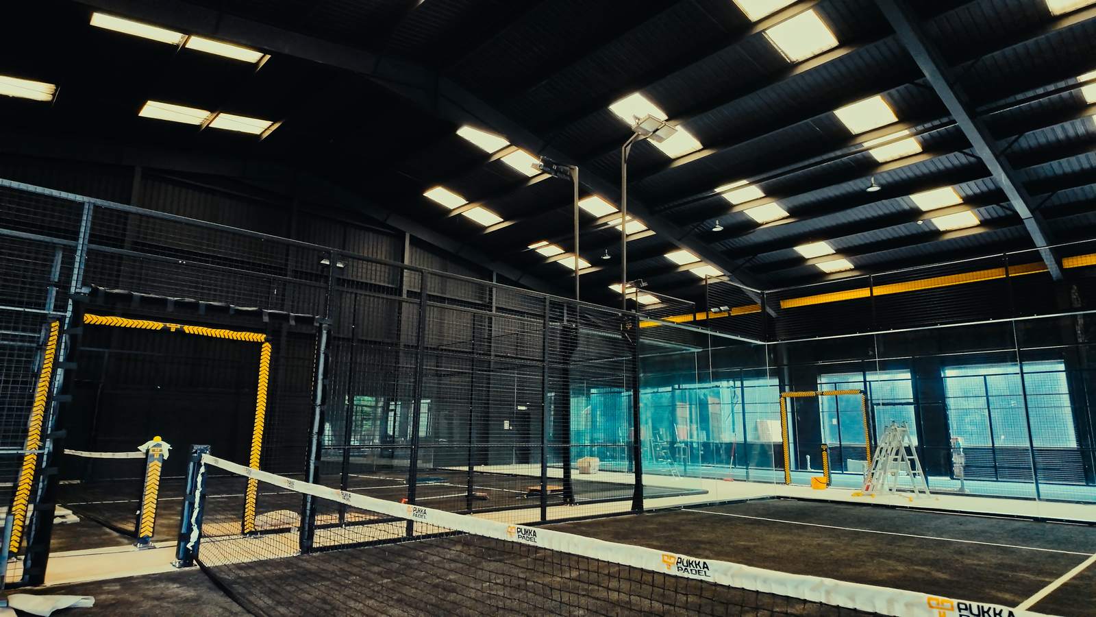 A multi-court indoor padel facility with black steel frames, glass walls and overhead lighting