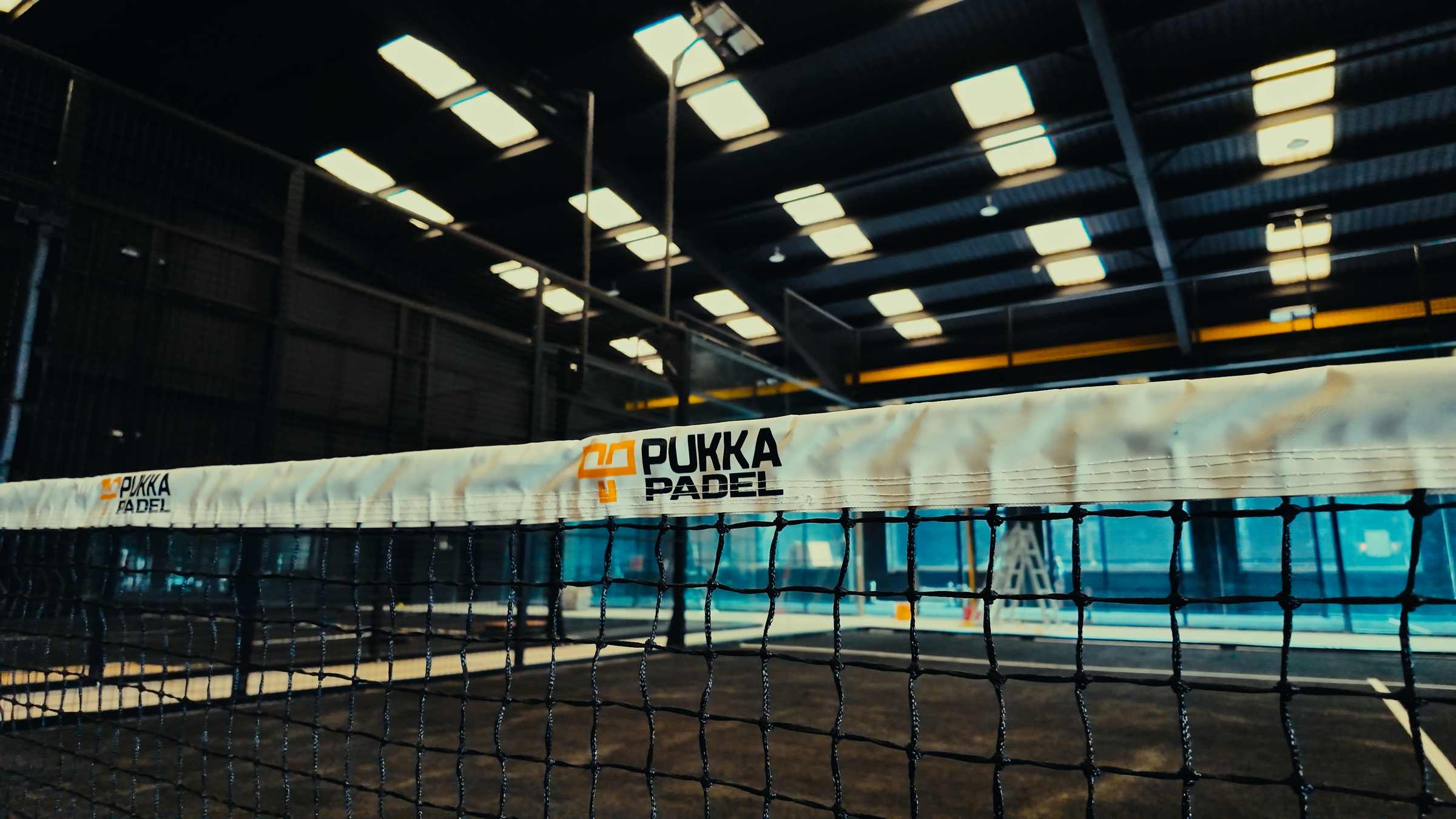 An indoor padel court with a Pukka Padel branded net, glass side walls and a lit steel roof overhead