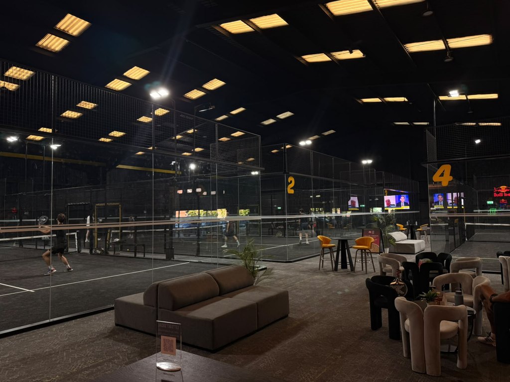 An indoor padel centre at night: numbered courts in play behind glass, with a fitted-out lounge and seating area in the foreground.