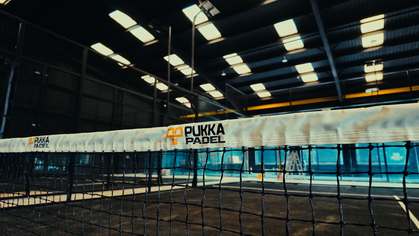 An indoor padel court with a Pukka Padel branded net, glass side walls and a lit steel roof overhead.